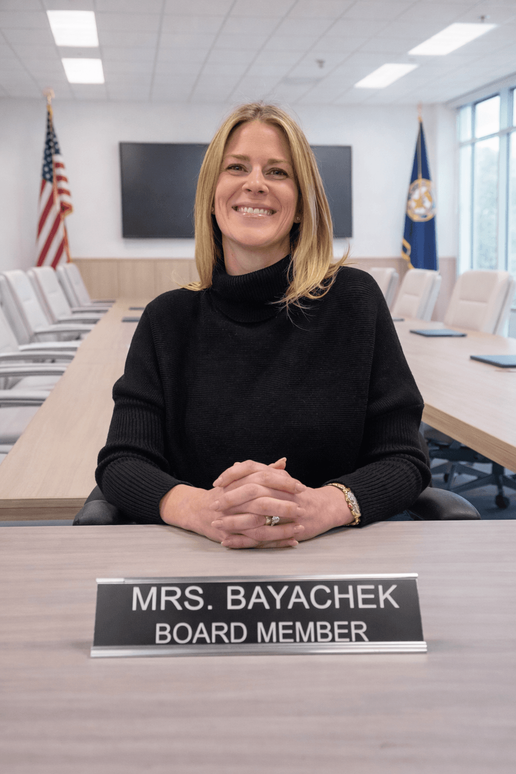 Smiling blonde woman in a black sweater sits behind a board member nameplate.