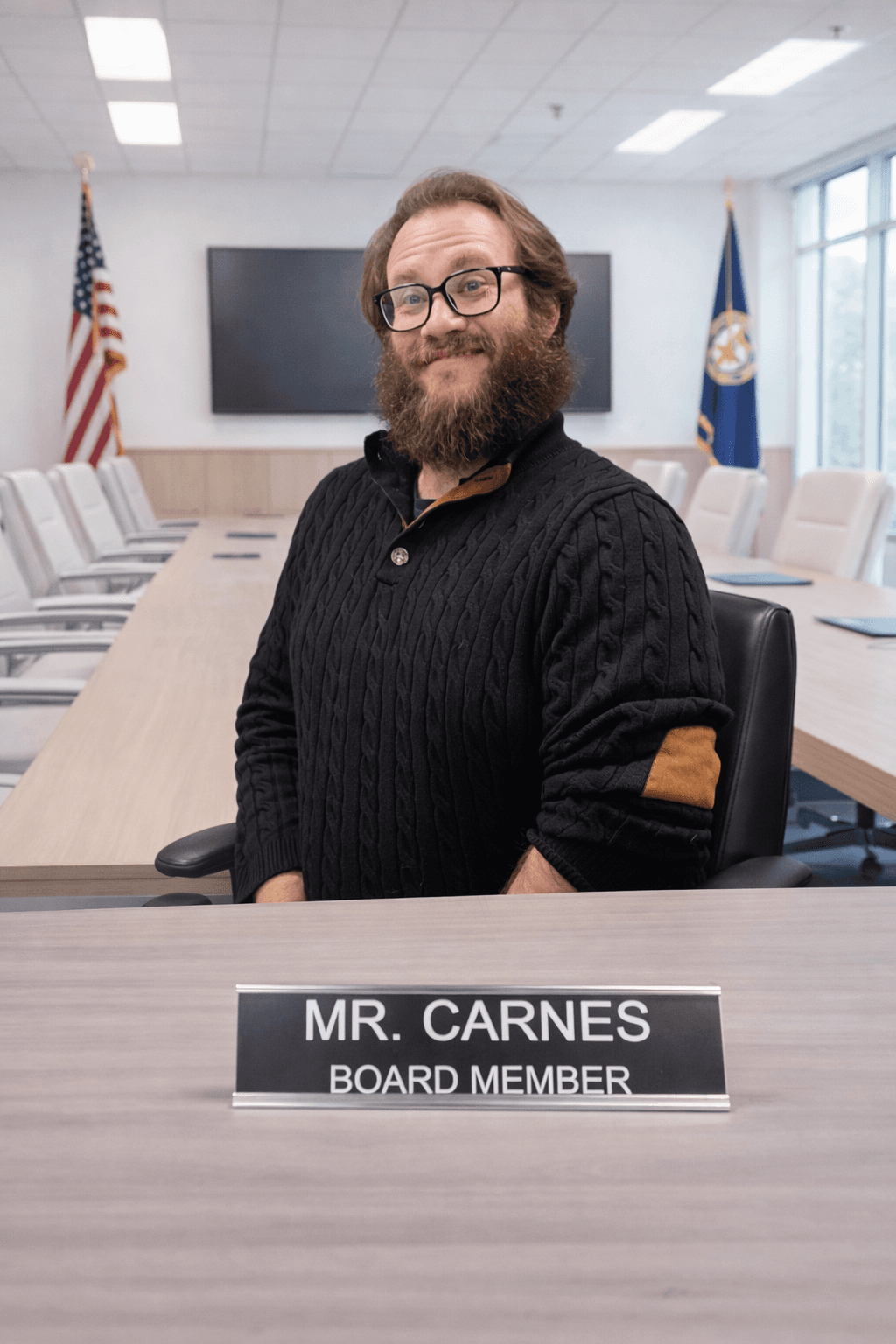 Bearded man in a black sweater sits behind a nameplate reading Mr. Carnes Board Member.