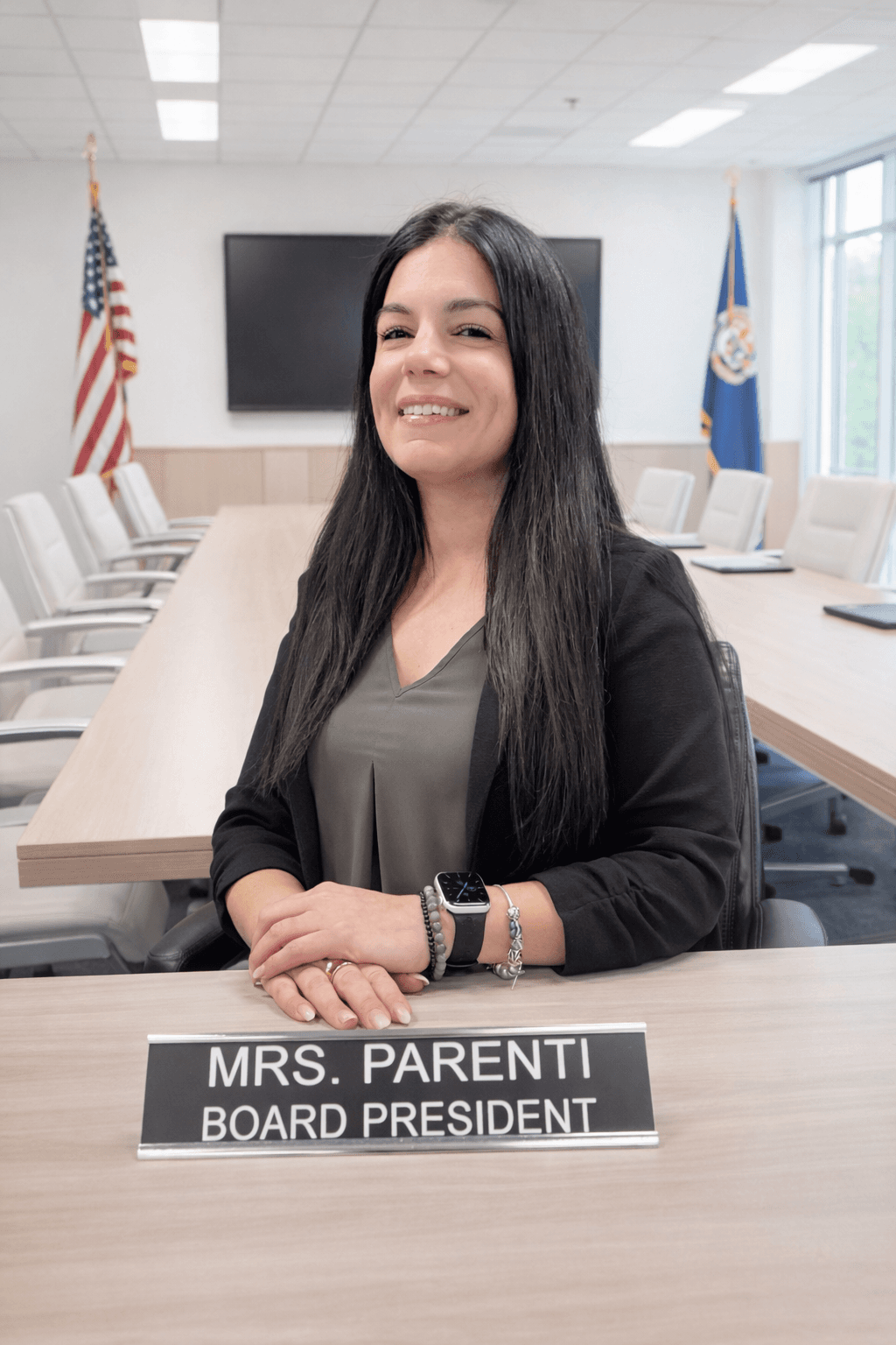 Smiling Board President Mrs. Parenti sits at a conference table with her nameplate and flags.