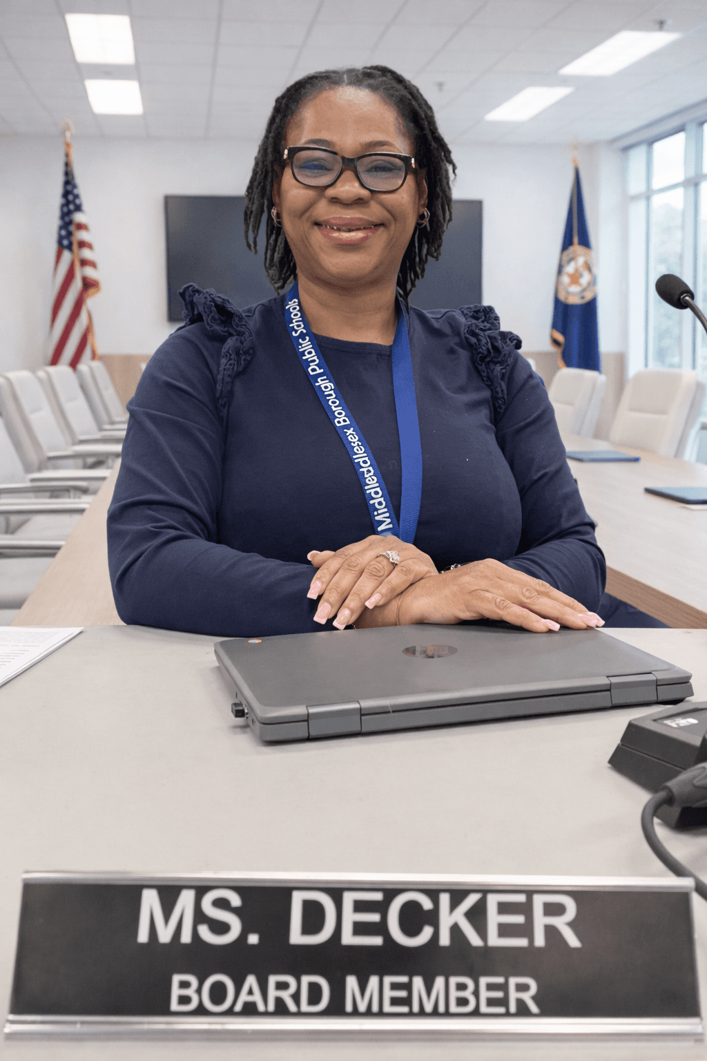Board member Ms. Decker smiles at a conference table with her nameplate and laptop.