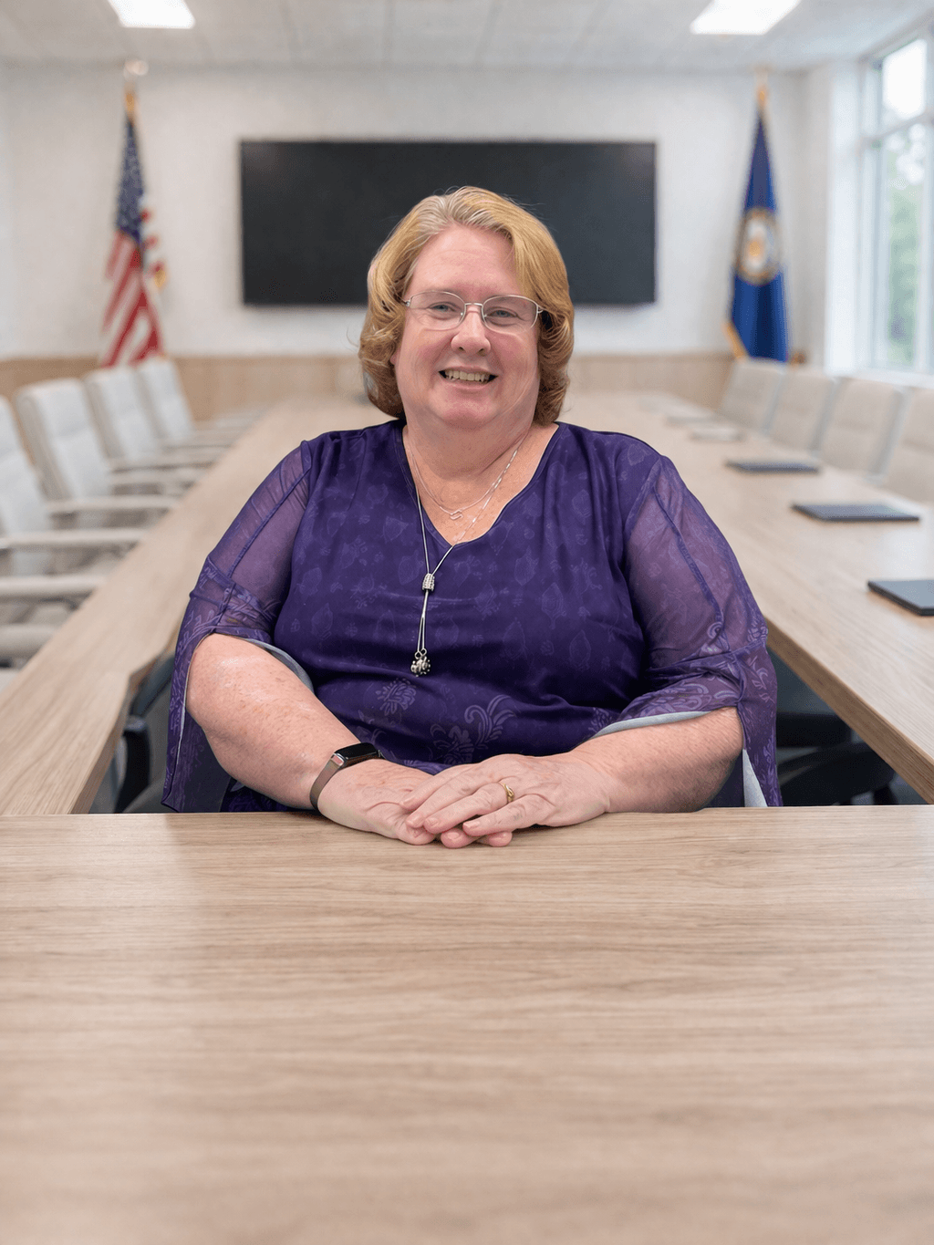 Smiling woman with glasses and purple top sits at a long boardroom table with flags.