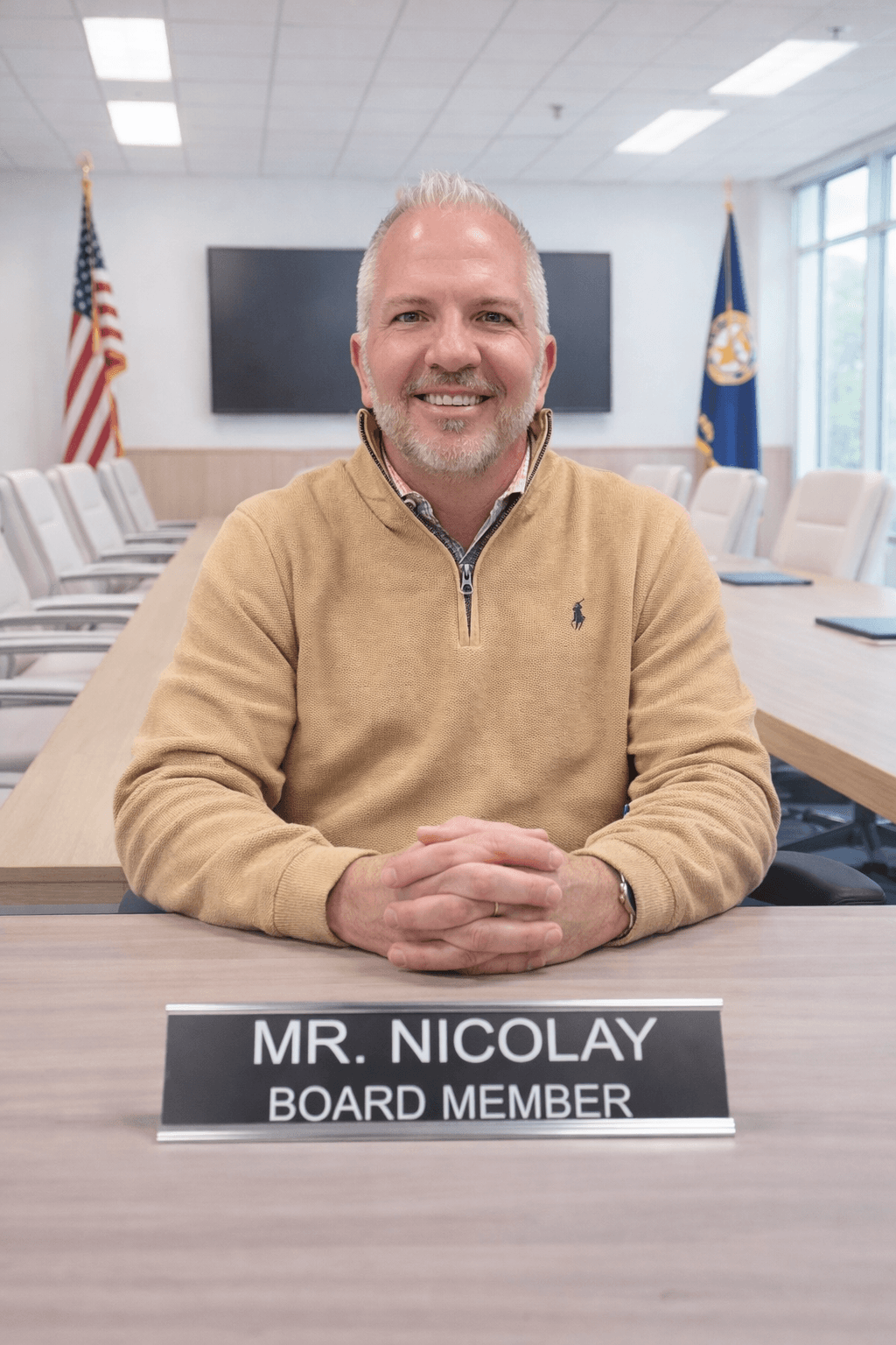 Board member Mr. Nicolay smiles at a conference table with flags in the background.