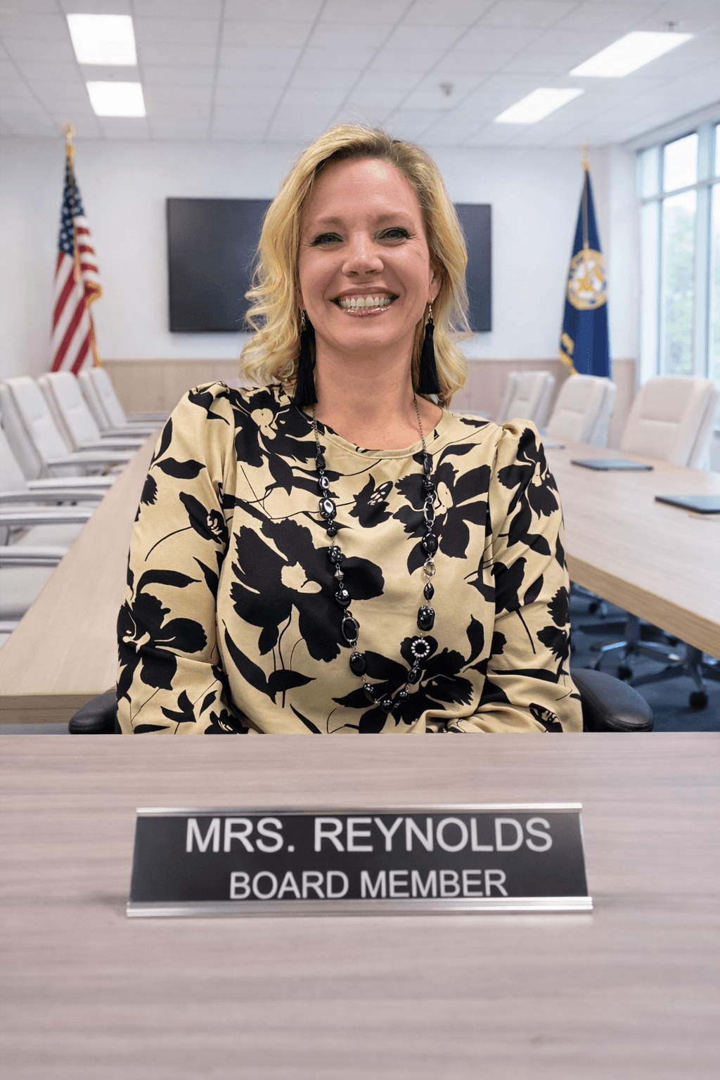 Smiling blonde board member Mrs. Reynolds sits at a conference table wearing a floral blouse.